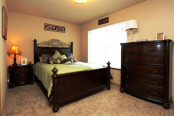 Apartment bedroom with queen set of furniture, bay window and carpeted floors.